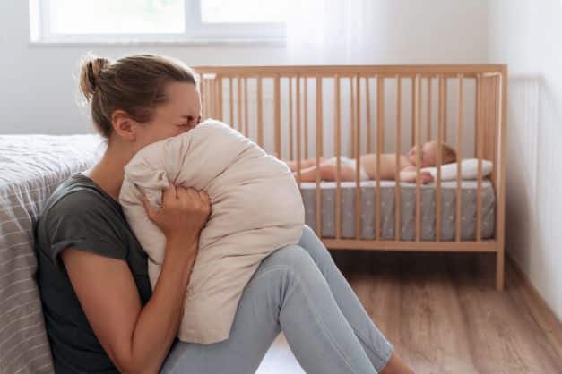 woman-sitting-on-the-floor-screeming-into-a-pillow-while-her-baby-is-in-the-crib