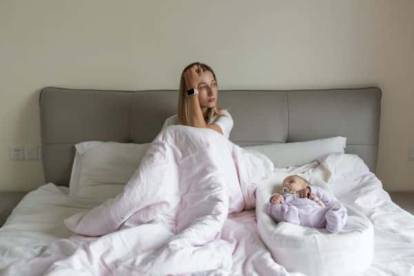 woman-sitting-in-bed-looking-overwhelmed-with-her-baby-lying-on-the-bed-in-a-sleeping-pillow